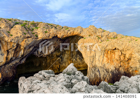 Sunlight above natural grotto entrance on a rocky coastline revealing the dark water within its cavernous depths Sunlight above natural grotto entrance on a rocky coastline revealing the dark water within its cavernous depths 141408091