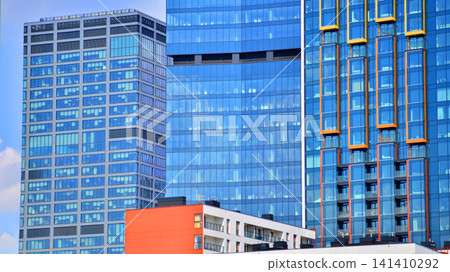 A low-rise residential building stands against a backdrop of tall glass buildings. An urban quarter of modern architecture. A low-rise residential building stands against a backdrop of tall glass buildings. An urban quarter of modern architecture. 141410292