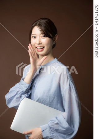 A smiling Japanese woman holding a laptop and calling out against a brown background; an image representing word-of-mouth, support, or business announcements/promotions. A smiling Japanese woman holding a laptop and calling out against a brown background; an image representing word-of-mouth, support, or business announcements/promotions. 141411270
