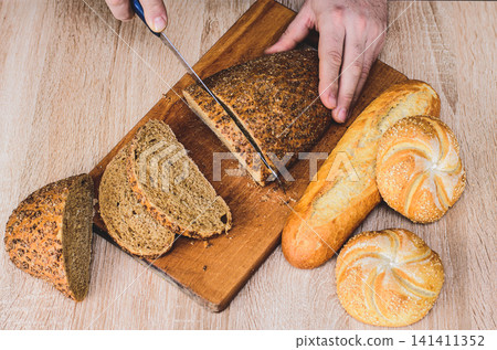 A man with a knife cuts bread on a wooden board. French baguettes. Different breeds on wooden background. A man with a knife cuts bread on a wooden board. French baguettes. Different breeds on wooden background. 141411352