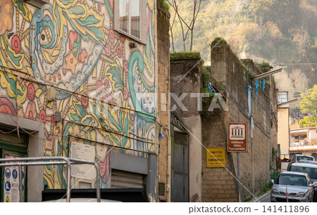 Naples, italy. December 30, 2024. Naples street scene featuring vibrant mural on building facade, alongside historic architecture and sign pointing to fontanelle cemetery Naples, italy. December 30, 2024. Naples street scene featuring vibrant mural on building facade, alongside historic architecture and sign pointing to fontanelle cemetery 141411896