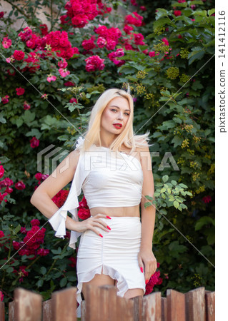 Adult blonde woman in a white outfit leans across a rustic wooden fence while looking at the camera. Red roses and dense foliage frame the outdoor portrait. Adult blonde woman in a white outfit leans across a rustic wooden fence while looking at the camera. Red roses and dense foliage frame the outdoor portrait. 141412116