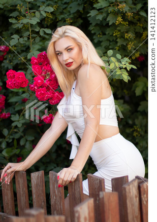 Adult blonde woman in a white outfit leans across a rustic wooden fence while looking at the camera. Red roses and dense foliage frame the outdoor portrait. Adult blonde woman in a white outfit leans across a rustic wooden fence while looking at the camera. Red roses and dense foliage frame the outdoor portrait. 141412123