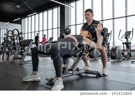 Personal trainer spotting woman performing dumbbell bench press in bright modern gym with cardio machines, large windows, natural light, focused strength training session Personal trainer spotting woman performing dumbbell bench press in bright modern gym with cardio machines, large windows, natural light, focused strength training session 141412663