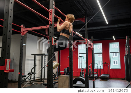 woman performing pull-ups in focused and determined side view of indoor gym with red and black equipment, strength training workout periodical resource for fitness industry use cases. woman performing pull-ups in focused and determined side view of indoor gym with red and black equipment, strength training workout periodical resource for fitness industry use cases. 141412673