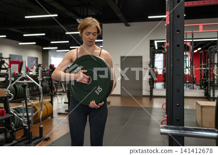 Female athlete loading heavy weight plate onto barbell in squat rack inside bright modern gym, focused expression, strong posture, and visible effort during strength training Female athlete loading heavy weight plate onto barbell in squat rack inside bright modern gym, focused expression, strong posture, and visible effort during strength training 141412678