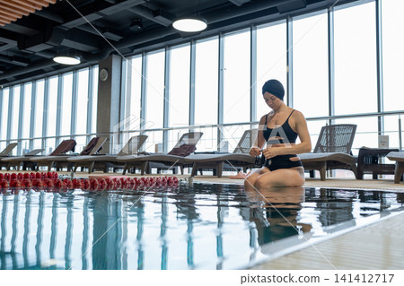 Young woman in black swimsuit sits beside indoor pool, feet in water, resting near lane divider inside modern swimming facility with wet hair, calm posture and natural light Young woman in black swimsuit sits beside indoor pool, feet in water, resting near lane divider inside modern swimming facility with wet hair, calm posture and natural light 141412717