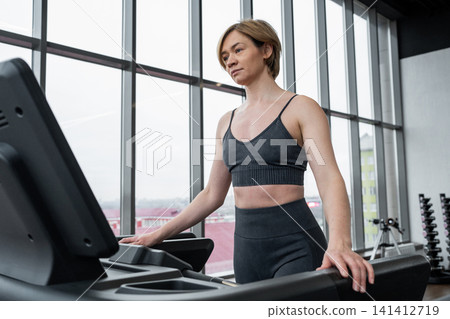 Young woman walking on treadmill in bright modern gym studio with floor-to-ceiling windows and city rooftops outside, surrounded by rows of cardio machines and soft daylight Young woman walking on treadmill in bright modern gym studio with floor-to-ceiling windows and city rooftops outside, surrounded by rows of cardio machines and soft daylight 141412719