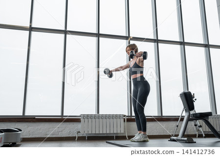 Athletic woman lifting dumbbells in lateral raise exercise inside bright modern gym with floor to ceiling windows and city skyline, focused posture and strong toned arms Athletic woman lifting dumbbells in lateral raise exercise inside bright modern gym with floor to ceiling windows and city skyline, focused posture and strong toned arms 141412736