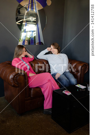 Women chatting and relaxing together in quiet corner of stylish cafe, one in pink suit. Cafe, quiet, chatting, pink suit, stylish. Women chatting and relaxing together in quiet corner of stylish cafe, one in pink suit. Cafe, quiet, chatting, pink suit, stylish. 141412788