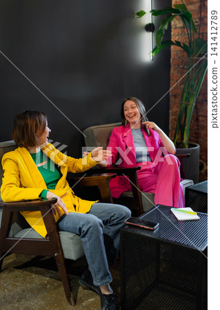 Cheerful women in yellow and pink suits smiling and chatting in cafe, friendly meeting over drinks. Cafe, chat, suits, smiles, drinks. Cheerful women in yellow and pink suits smiling and chatting in cafe, friendly meeting over drinks. Cafe, chat, suits, smiles, drinks. 141412789