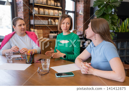 Three women meeting at cozy cafe table with notepads and coffee, smiling and planning together. Cafe, women, planning, coffee, notepads. Three women meeting at cozy cafe table with notepads and coffee, smiling and planning together. Cafe, women, planning, coffee, notepads. 141412875