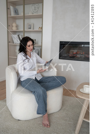 Relaxing at home, woman reading magazine on cozy white chair, drinking morning coffee, enjoying leisure time. Lifestyle, relaxation, comfort, tranquility, indoors Relaxing at home, woman reading magazine on cozy white chair, drinking morning coffee, enjoying leisure time. Lifestyle, relaxation, comfort, tranquility, indoors 141412893