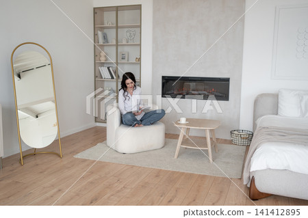 Relaxing at home, woman reading magazine on cozy white chair, drinking morning coffee, enjoying leisure time. Lifestyle, relaxation, comfort, tranquility, indoors Relaxing at home, woman reading magazine on cozy white chair, drinking morning coffee, enjoying leisure time. Lifestyle, relaxation, comfort, tranquility, indoors 141412895