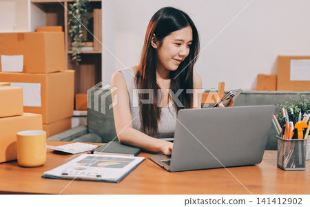 Startup SME small business entrepreneur of freelance Asian woman wearing apron using laptop and box to receive and review orders online to prepare to pack sell to customers, online sme business ideas. Startup SME small business entrepreneur of freelance Asian woman wearing apron using laptop and box to receive and review orders online to prepare to pack sell to customers, online sme business ideas. 141412902