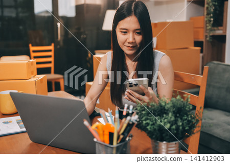 Startup SME small business entrepreneur of freelance Asian woman wearing apron using laptop and box to receive and review orders online to prepare to pack sell to customers, online sme business ideas. 141412903