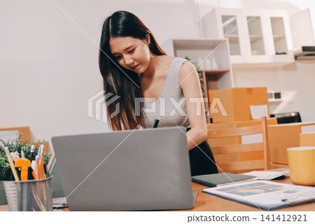 Startup SME small business entrepreneur of freelance Asian woman wearing apron using laptop and box to receive and review orders online to prepare to pack sell to customers, online sme business ideas. Startup SME small business entrepreneur of freelance Asian woman wearing apron using laptop and box to receive and review orders online to prepare to pack sell to customers, online sme business ideas. 141412921