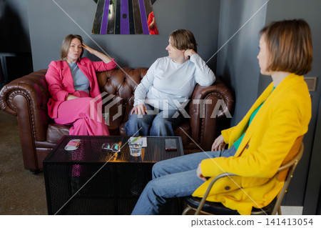 Three women enjoying coffee together and talking in fashionable city cafe, brown sofa and wood decor. Cafe, coffee, talking, wood, city, sofa. Three women enjoying coffee together and talking in fashionable city cafe, brown sofa and wood decor. Cafe, coffee, talking, wood, city, sofa. 141413054