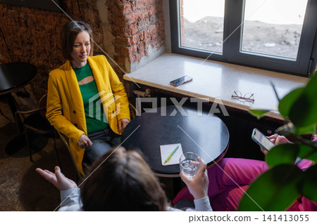 Top-down view of businesslike women in yellow and white meeting by window in cozy cafe nook. Cafe, top down, business, yellow, window. Top-down view of businesslike women in yellow and white meeting by window in cozy cafe nook. Cafe, top down, business, yellow, window. 141413055