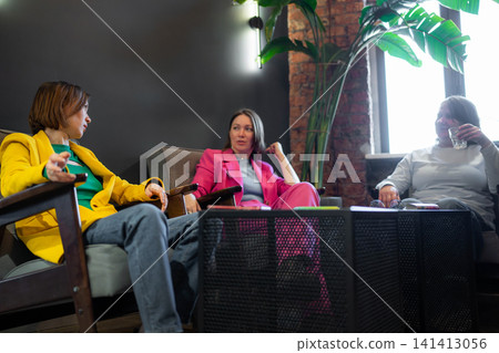 Women in yellow and pink jackets chatting and laughing with friend at black table in cafe. Cafe, jackets, chatting, laughing, table. Women in yellow and pink jackets chatting and laughing with friend at black table in cafe. Cafe, jackets, chatting, laughing, table. 141413056