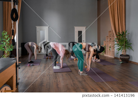 Young caucasian woman doing roll down exercise in a pilates bright studio. Their workout focuses on core strength and flexibility in a modern fitness environment Young caucasian woman doing roll down exercise in a pilates bright studio. Their workout focuses on core strength and flexibility in a modern fitness environment 141413196