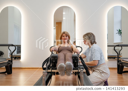 Sporty mature woman practicing pilates abs routine on reformer machine in daylight studio with trainer. Pilates abs, reformer machine, sporty, studio, routine Sporty mature woman practicing pilates abs routine on reformer machine in daylight studio with trainer. Pilates abs, reformer machine, sporty, studio, routine 141413293