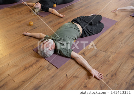 Senior gray-haired adult man doing pilates exercises for back and joint on a mat. A trainer helps him gently recover Senior gray-haired adult man doing pilates exercises for back and joint on a mat. A trainer helps him gently recover 141413331
