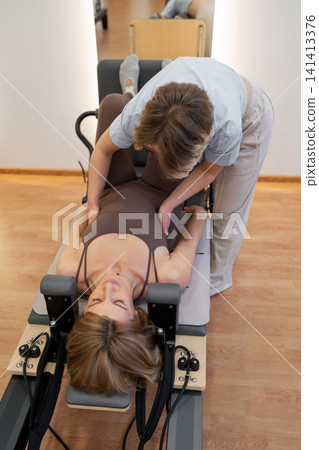 Sporty mature woman practicing pilates abs routine on reformer machine in daylight studio with trainer. Pilates abs, reformer machine, sporty, studio, routine Sporty mature woman practicing pilates abs routine on reformer machine in daylight studio with trainer. Pilates abs, reformer machine, sporty, studio, routine 141413376