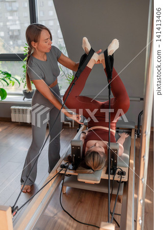 Woman holding high leg stretch with instructor's assistance on pilates reformer in neutral studio. High leg stretch, pilates reformer, assistance, instructor, studio. Woman holding high leg stretch with instructor's assistance on pilates reformer in neutral studio. High leg stretch, pilates reformer, assistance, instructor, studio. 141413406