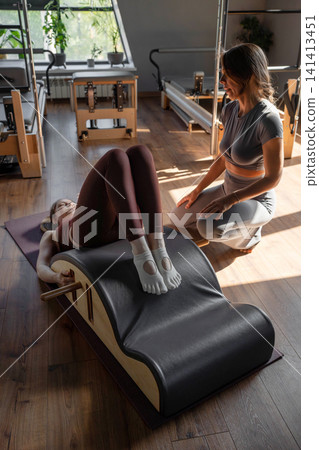 Woman doing exercises on back spine corrector with trainer. Reformer pilates studio machine for fitness workouts in gym. Fit, healthy and strong authentical body. Fitness concept Woman doing exercises on back spine corrector with trainer. Reformer pilates studio machine for fitness workouts in gym. Fit, healthy and strong authentical body. Fitness concept 141413451