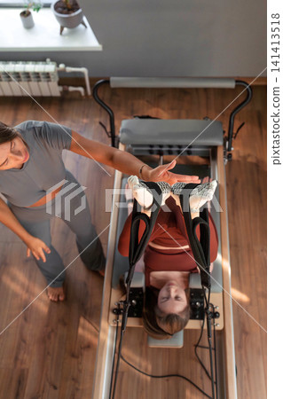 Woman holding high leg stretch with instructor's assistance on pilates reformer in neutral studio. High leg stretch, pilates reformer, assistance, instructor, studio. Woman holding high leg stretch with instructor's assistance on pilates reformer in neutral studio. High leg stretch, pilates reformer, assistance, instructor, studio. 141413518