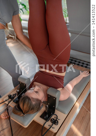 Woman holding high leg stretch with instructor's assistance on pilates reformer in neutral studio. High leg stretch, pilates reformer, assistance, instructor, studio. Woman holding high leg stretch with instructor's assistance on pilates reformer in neutral studio. High leg stretch, pilates reformer, assistance, instructor, studio. 141413522