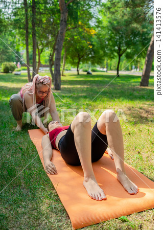 Fitness instructor helping student do knee lifts on orange mat during outdoor park exercise class. Knee lifts, fitness, instructor, orange mat, outdoor, park. 141413576