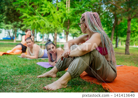 Ladies relaxing after yoga session sitting on orange mat together in city park, chatting and laughing. Relaxing, yoga, mat, chatting, park, after session. 141413640