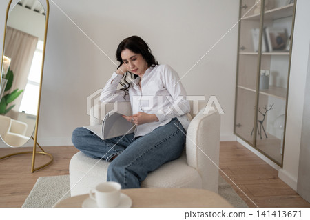 Relaxing at home, woman reading magazine on cozy white chair, drinking morning coffee, enjoying leisure time. Lifestyle, relaxation, comfort, tranquility, indoors Relaxing at home, woman reading magazine on cozy white chair, drinking morning coffee, enjoying leisure time. Lifestyle, relaxation, comfort, tranquility, indoors 141413671