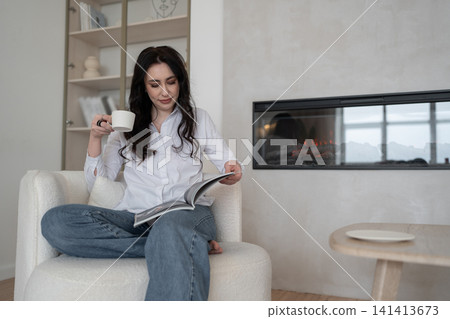 Relaxing at home, woman reading magazine on cozy white chair, drinking morning coffee, enjoying leisure time. Lifestyle, relaxation, comfort, tranquility, indoors Relaxing at home, woman reading magazine on cozy white chair, drinking morning coffee, enjoying leisure time. Lifestyle, relaxation, comfort, tranquility, indoors 141413673