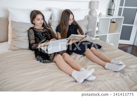 Friends laying on bed working on school assignments with notebooks and computer, smiling. Assignments, bed, friends, notebooks, school, smiling. Friends laying on bed working on school assignments with notebooks and computer, smiling. Assignments, bed, friends, notebooks, school, smiling. 141413775