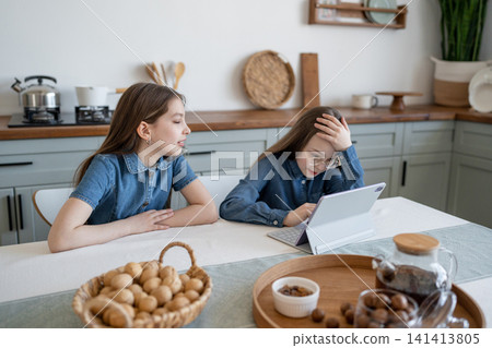 Frustrated girl leaning head on hand while doing homework at wooden breakfast table. Frustrated, homework, wood table, girl, breakfast. Frustrated girl leaning head on hand while doing homework at wooden breakfast table. Frustrated, homework, wood table, girl, breakfast. 141413805