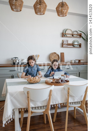 Two girls sitting at round kitchen table with notepads and eating pastries together in cozy home. Notepads, table, eating, pastries, kitchen, girls. Two girls sitting at round kitchen table with notepads and eating pastries together in cozy home. Notepads, table, eating, pastries, kitchen, girls. 141413820