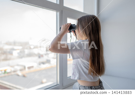 Girl with long hair looking through binoculars at window on bright day in cozy room. Binoculars, window, girl, bright, room, curiosity. Girl with long hair looking through binoculars at window on bright day in cozy room. Binoculars, window, girl, bright, room, curiosity. 141413883