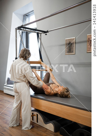 Pilates mature woman doing back strengthening exercises on in pilates studio. Balanced Body Cadillac machine for fitness workouts in gym. Fit, healthy and strong authentical body. Fitness concept. Pilates mature woman doing back strengthening exercises on in pilates studio. Balanced Body Cadillac machine for fitness workouts in gym. Fit, healthy and strong authentical body. Fitness concept. 141413918