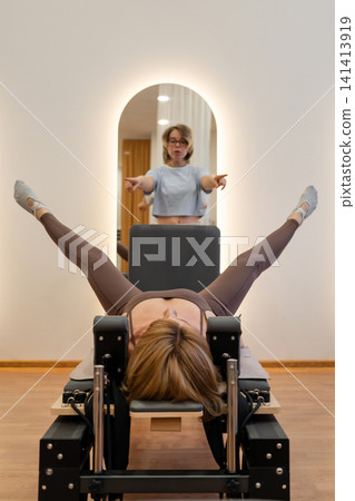 Woman training pilates with trainer on the reformer bed. Reformer pilates studio machine for fitness workouts in gym. Fit, healthy and strong authentical body. Fitness Woman training pilates with trainer on the reformer bed. Reformer pilates studio machine for fitness workouts in gym. Fit, healthy and strong authentical body. Fitness 141413919