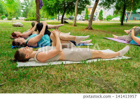 Group of women performing yoga pose lying on mats with hands around legs in city park. Yoga pose, legs, mats, city park, group, women. Group of women performing yoga pose lying on mats with hands around legs in city park. Yoga pose, legs, mats, city park, group, women. 141413957