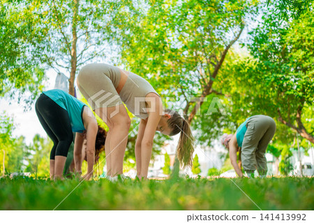Women in fitness clothes practicing forward bend on mats in sunny park group yoga class. Forward bend, yoga, park, group, women. 141413992