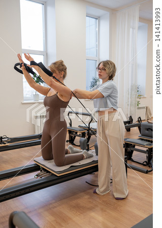 A woman in activewear does pilates exercises for the chest and back muscles with a reformer machine in a sunlite studio, committed to her workout routine A woman in activewear does pilates exercises for the chest and back muscles with a reformer machine in a sunlite studio, committed to her workout routine 141413993
