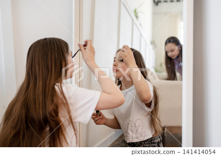 Girls applying mascara and lipstick happily in front of mirror in airy white bedroom. Blush, lipstick, mirror, girls, happy, white, bedroom. Girls applying mascara and lipstick happily in front of mirror in airy white bedroom. Blush, lipstick, mirror, girls, happy, white, bedroom. 141414054
