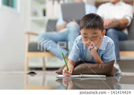 Asian boy doing homework on floor with parents working in background 141414424