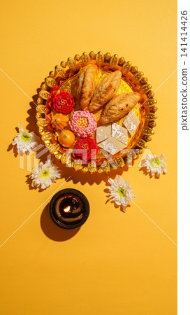 Diwali festive thali with traditional Indian sweets and lit diya on yellow background 141414426