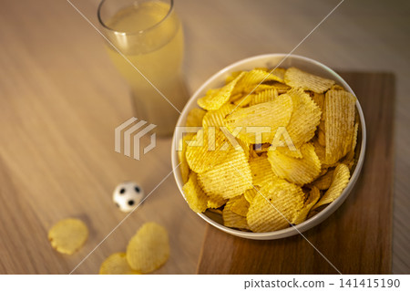 A glass of beer and potato chips snack on wooden board during soccer match, highlighting comforting food for big football events. 141415190