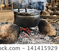 A close-up of an old aluminium pot placed over a rustic wood campfire supported by stones. Showing glowing embers and ashes, representing rural lifestyle and traditional outdoor cooking. 141415929